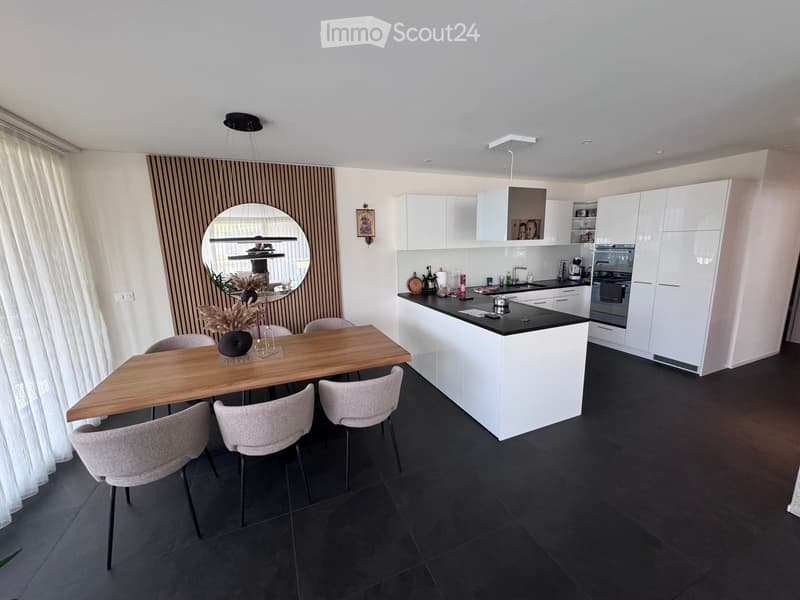 Modern kitchen with white cabinets, black countertops, a wooden dining table, and four chairs. Features a large island with sink and appliances, a round mirror, and a hanging light fixture.