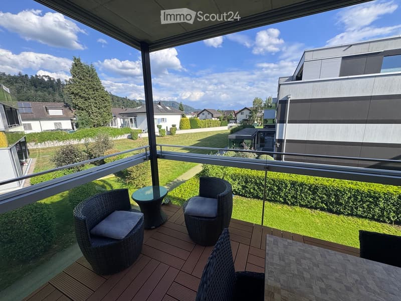 Balcony with wooden decking, glass railing, and outdoor furniture including a table and chairs, overlooking a green lawn and residential area.