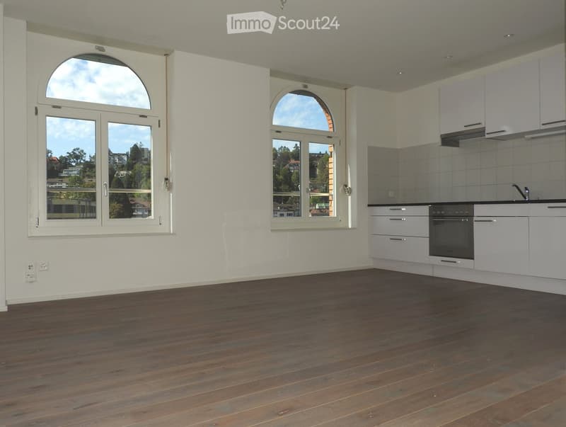 Empty room with wooden flooring, white walls, large windows with arched tops, and a small kitchen area with cabinets and an oven.