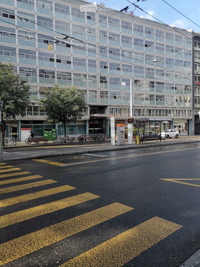 Modern multi-story building with large windows and a glass facade, located on a street corner with a bus stop, bicycles, and a pedestrian crossing.