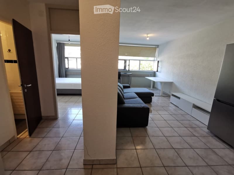 A modern, open-plan living and sleeping area with tiled flooring, a grey sectional sofa, a white coffee table, a refrigerator, and a bed visible through an open doorway.