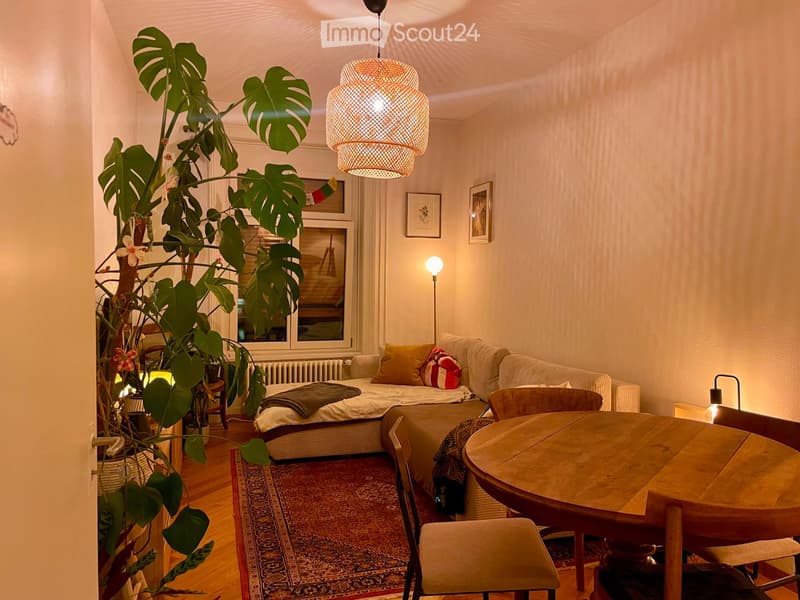 A cozy living room with a sofa, a small bed, a wooden table, chairs, a large leafy plant, and warm lighting.