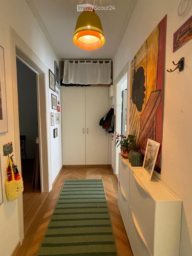 Hallway with wooden flooring, white walls, a hanging lamp, a green and white striped rug, a white cabinet, and various wall decorations including framed pictures and a large poster.