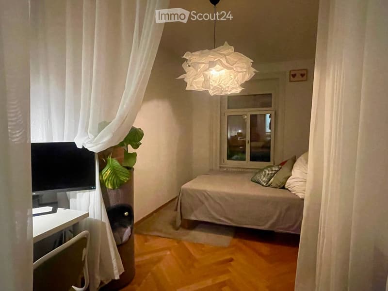 Cozy bedroom with a single bed, desk, chair, and a large window with white curtains. A potted plant is placed near the window, and a modern lamp hangs from the ceiling.