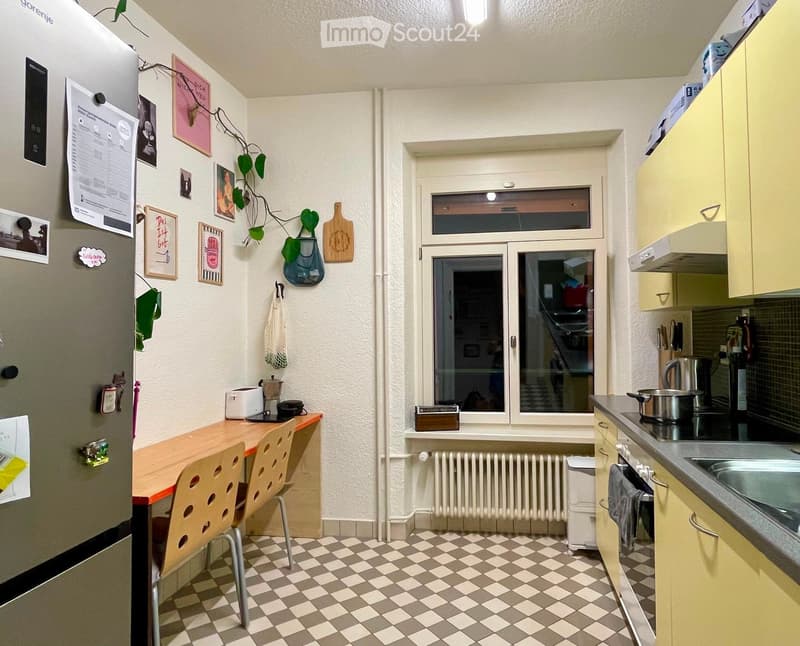 The kitchen features a checkered floor, a wooden table with two chairs, a refrigerator with various magnets and pictures, a window with a view, and built-in cabinets with a sink and stove.