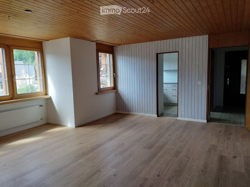 Unfurnished Room. Empty room with wooden flooring, white walls, and large windows. The room has a wooden ceiling and a radiator under one window. A doorway leads to another room with white cabinets.