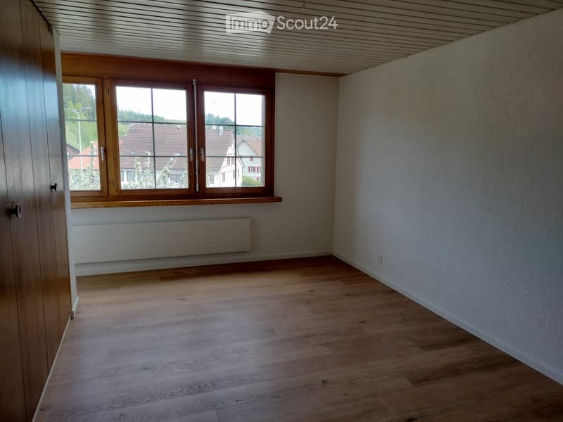 Unfurnished Room. Empty room with wooden flooring, white walls, large window with wooden frame, radiator, and a door visible on the left side.