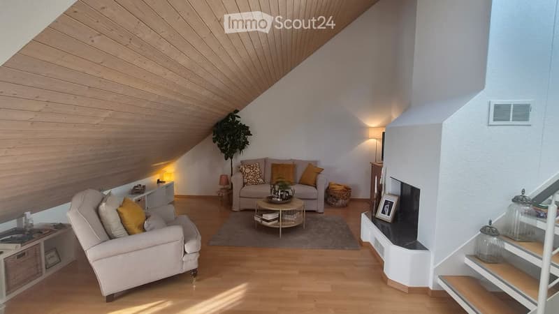 Cozy living room with wooden floors, slanted wooden ceiling, white walls, and warm lighting. Features a sofa, armchair, coffee table, side table, and decorative items.