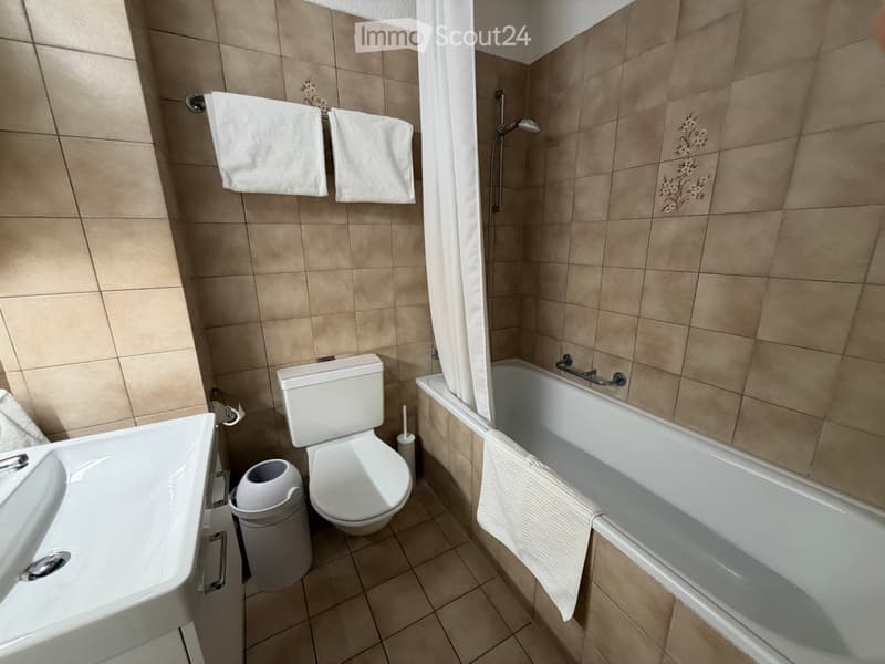 Bathroom with beige tiled walls and floor, featuring a toilet, bathtub, sink, and towels hanging on the wall.