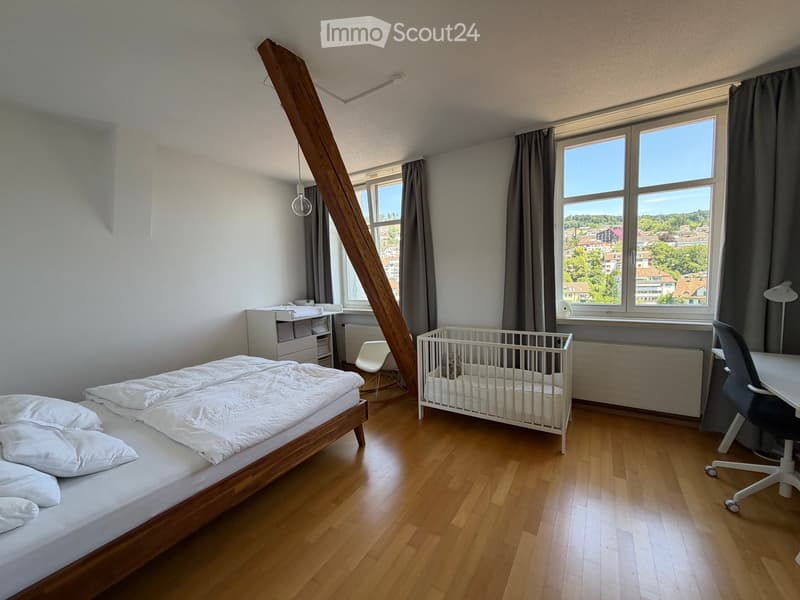 Bright bedroom with wooden flooring, large windows offering a scenic view, a double bed with white bedding, a baby crib, a small table with a chair, and a cabinet.