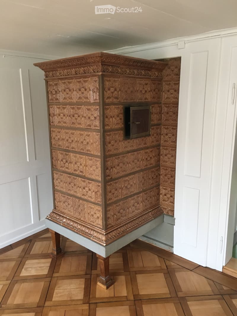 Ornate wooden fireplace with intricate carvings and a decorative mantelpiece, set against light grey walls and a wooden parquet floor.