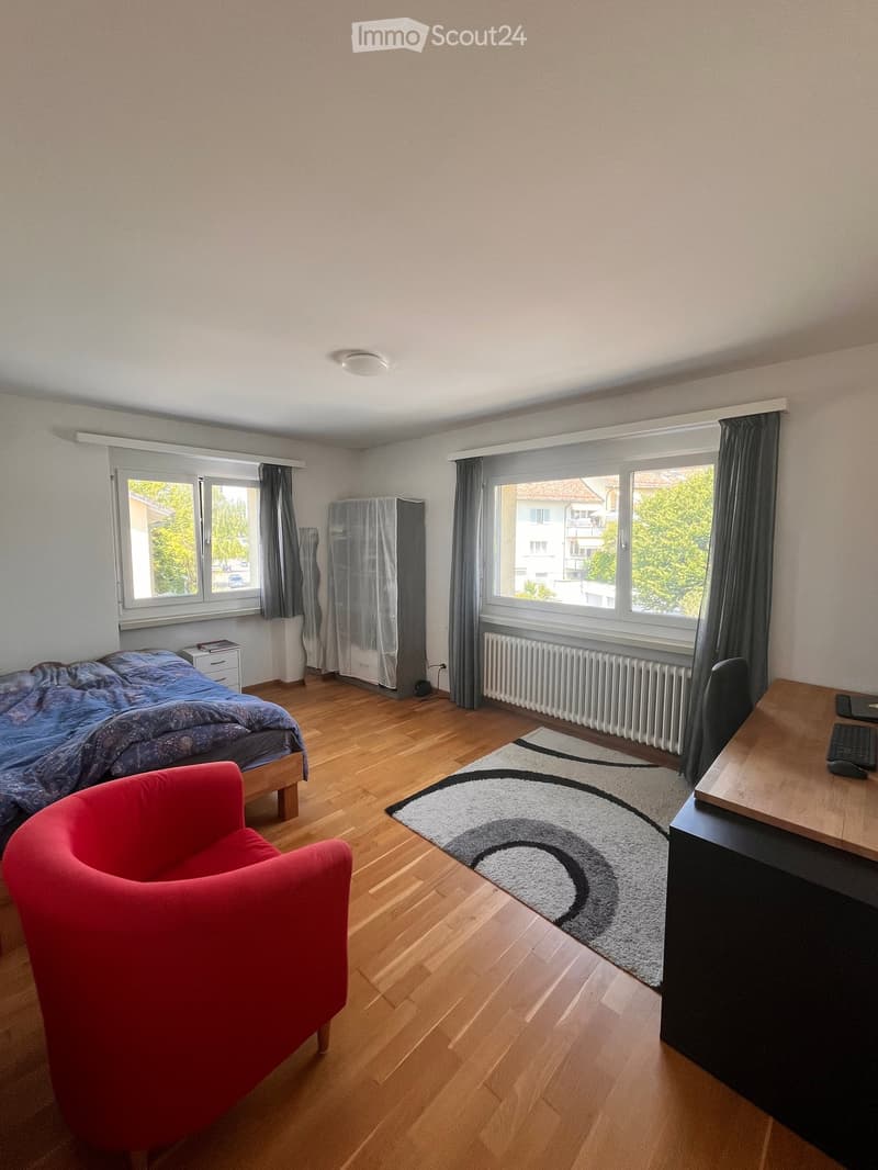 Bright bedroom with wooden flooring, large windows, a bed with blue bedding, a red armchair, a desk with a keyboard, and a patterned rug.