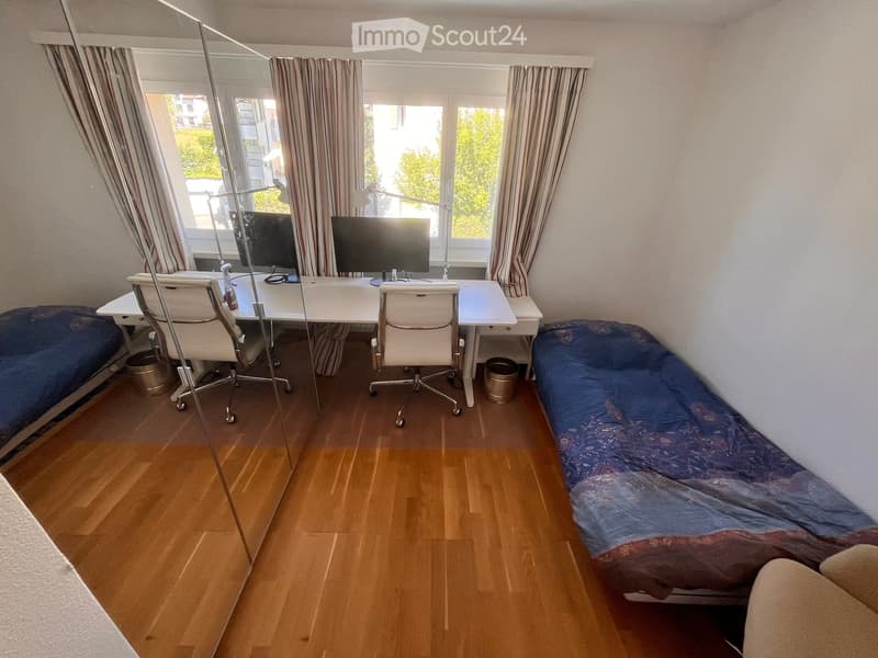 A furnished room with a desk, two chairs, a screen, and a sofa, featuring wooden flooring and large windows with curtains.