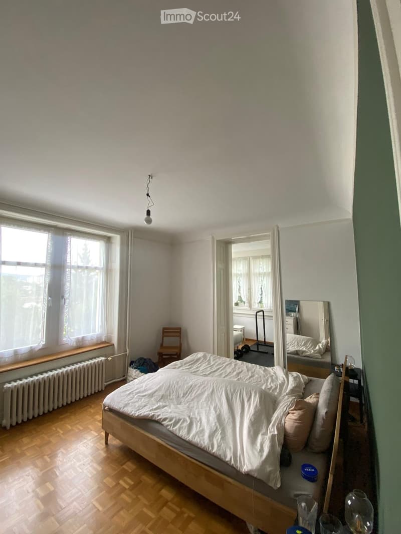 A well-lit bedroom with wooden flooring, a large bed with white bedding, a nightstand with a lamp, a mirror, a small wooden chair, and a radiator under a window.