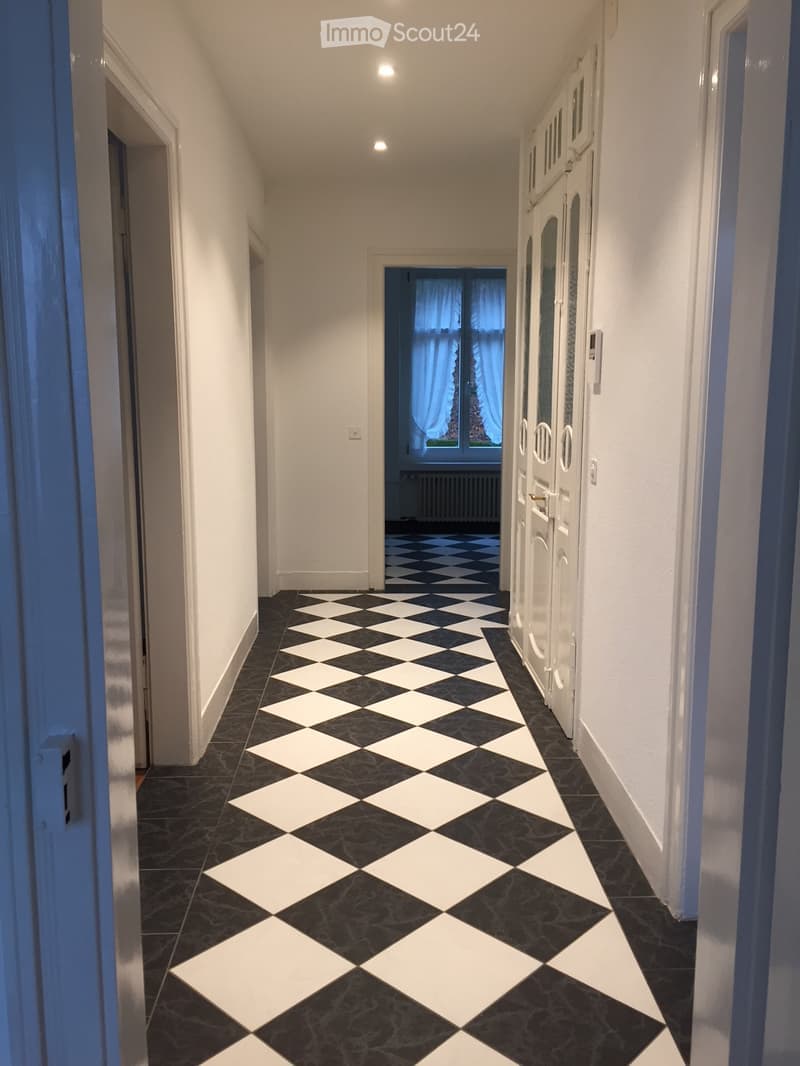 A long, narrow hallway with white walls, black and white checkered flooring, and several doors on the right side. The hallway is well-lit with recessed ceiling lights and has a window at the far end with blue curtains.