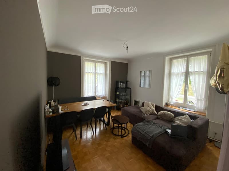 A modern living and dining area with a grey sofa, wooden dining table, chairs, a small round stool, and a laptop on the sofa. The room features large windows with white curtains and a wooden floor.