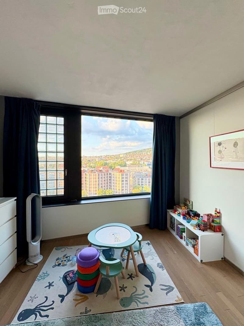 Brightly lit room with large windows offering a city view, featuring a small round table with chairs, a shelf with toys, a fan, and a colorful rug with sea animal designs.