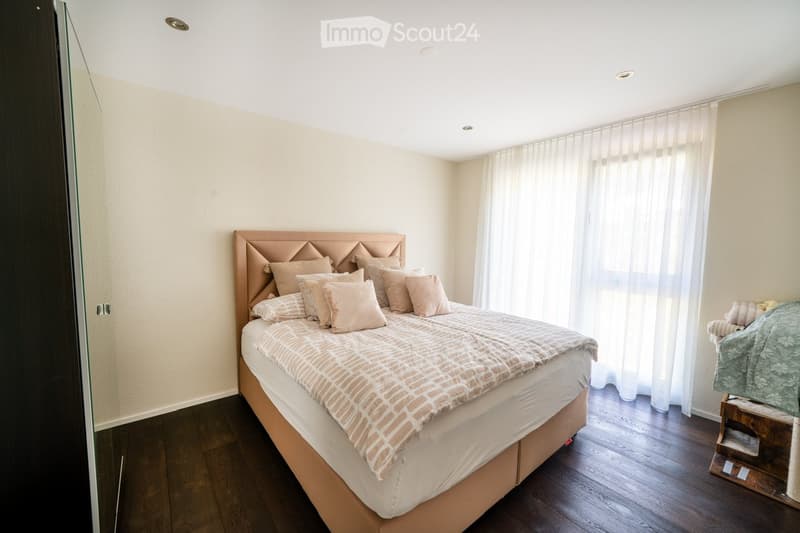 large bed, multiple pillows, beige duvet, window with white curtains, wooden floor