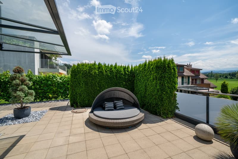 Patio with large sunbed, potted plant, round stone, hedges, and building in background