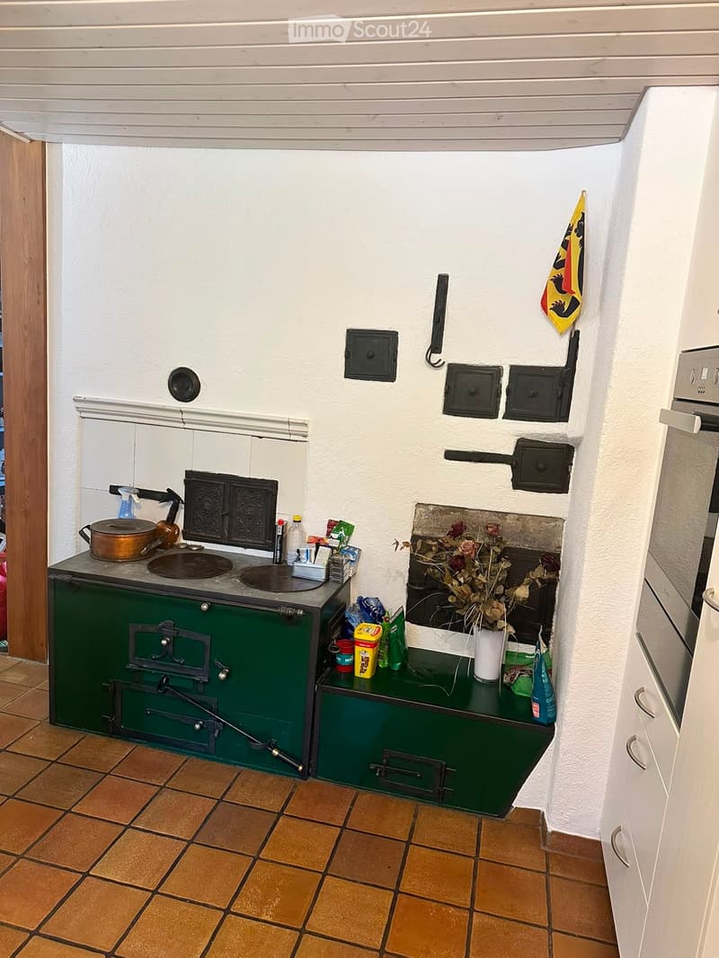 A rustic kitchen with a green stove, sink, and various kitchen items on the counter, including a vase with flowers and several bottles and cups.