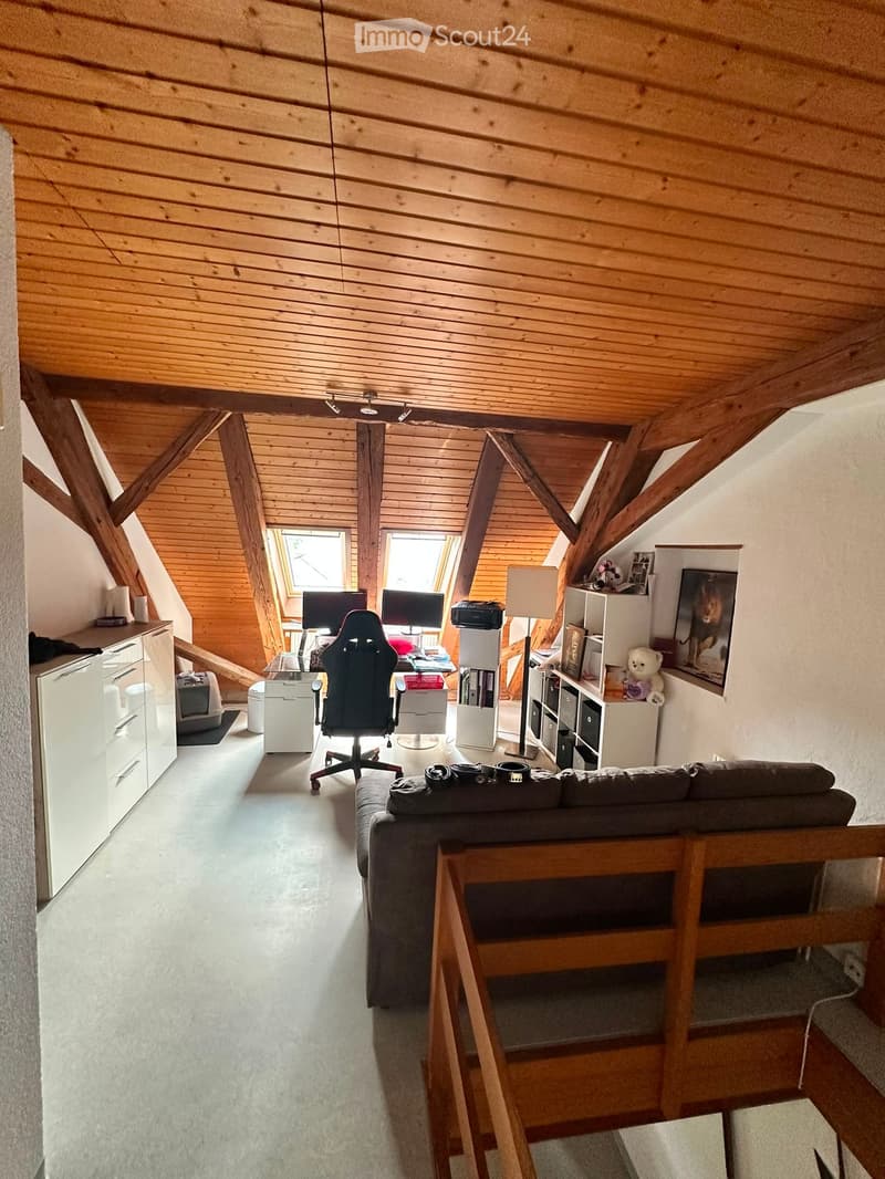 A cozy attic room with wooden beams and slanted ceilings, featuring a sofa, a gaming chair, a table with two screens, shelves, and a lamp.