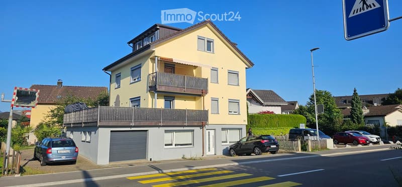 Two-story yellow house with a garage, balconies, and multiple windows. Several cars are parked in front.