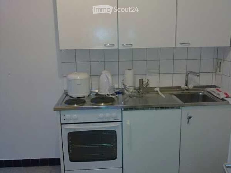 A compact kitchen with white cabinets, a small electric stove, a sink with a faucet, and various small appliances on the counter.