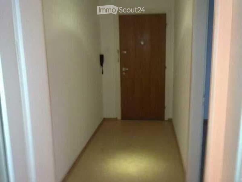 A narrow hallway with a wooden door at the end, beige walls, and a light-colored carpeted floor.