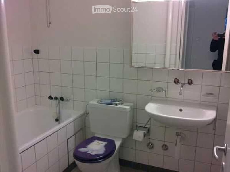 A small bathroom with white tiled walls and floor, featuring a toilet with a purple seat cover, a sink, a bathtub, and a mirror above the sink.
