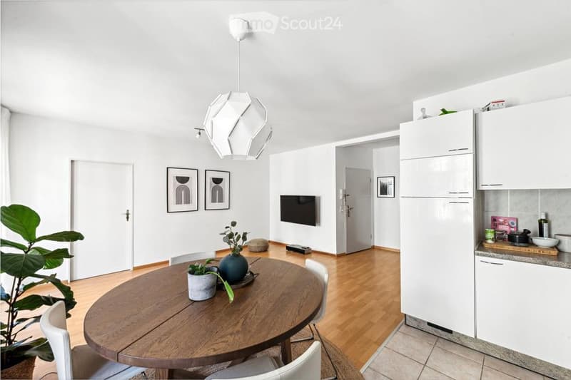 A modern, open-plan living and dining area with a round wooden dining table, white chairs, a hanging geometric light fixture, a television mounted on the wall, and a small kitchen area with white cabinets and a refrigerator.