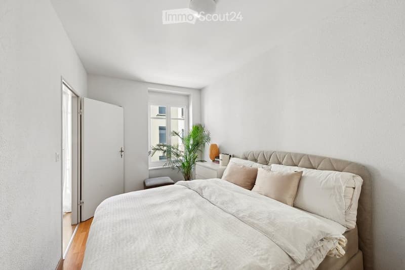 Bright bedroom with white walls, wooden floor, large window, bed with beige bedding, nightstand, plant, and lamp.