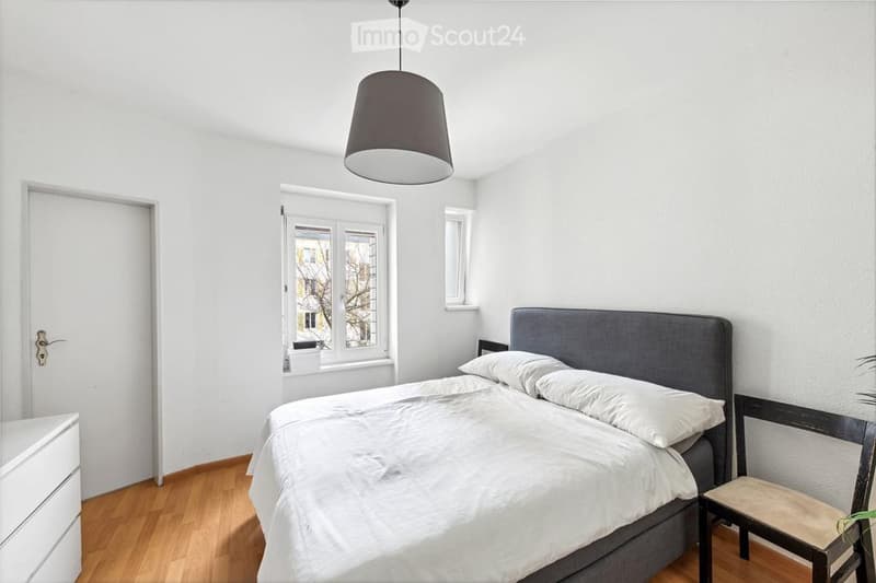 Bright bedroom with wooden flooring, white walls, a grey upholstered bed, a nightstand, a chair, and a hanging lamp.