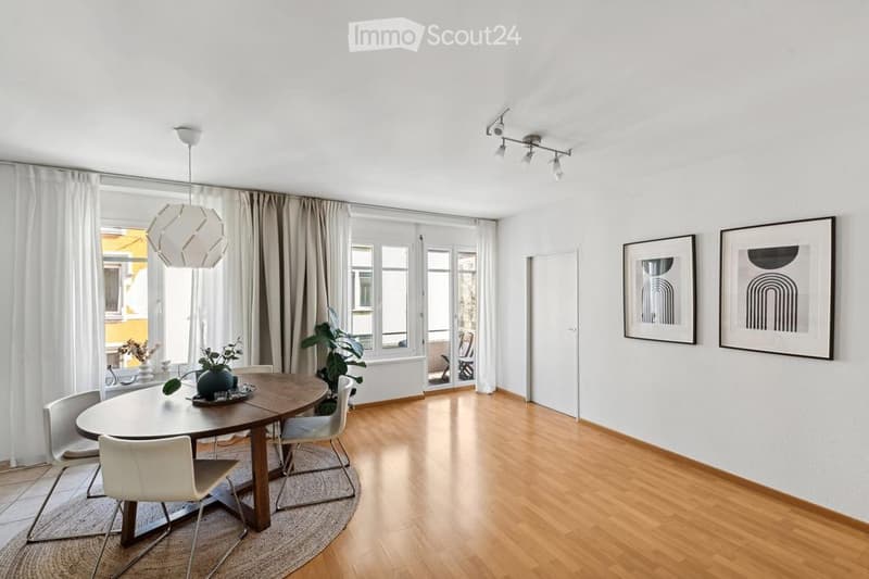 Bright dining area with a round wooden table, white chairs, pendant light, framed artwork, and plants near large windows with curtains.