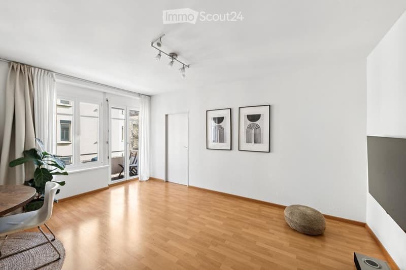 Bright, minimalist living space with wooden flooring, large windows, and a sliding glass door leading to a balcony. Includes a small table, chairs, a round stool, a potted plant, and two framed abstract artworks on the wall.