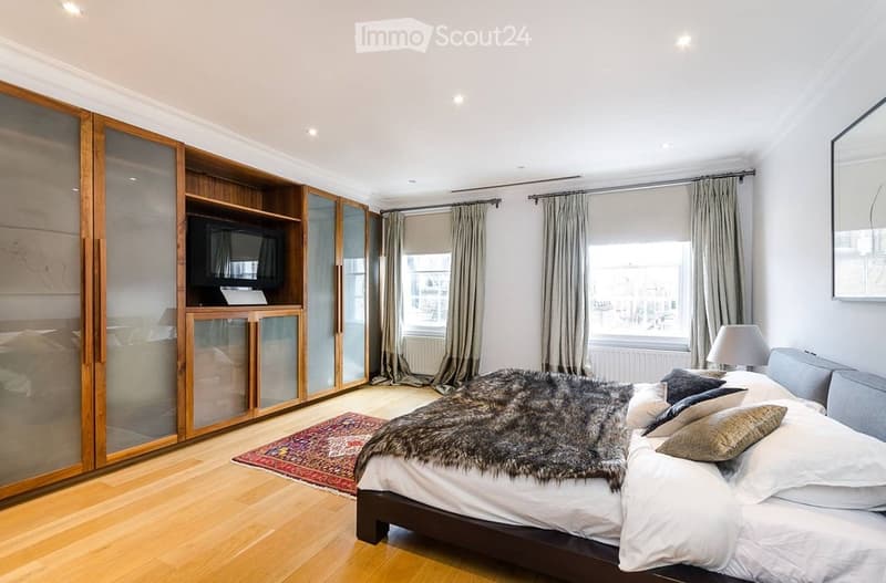 Modern bedroom with a large bed, wooden wardrobe, large mirror, and a television. The room features a plush fur throw on the bed, a patterned rug, and large windows with curtains.