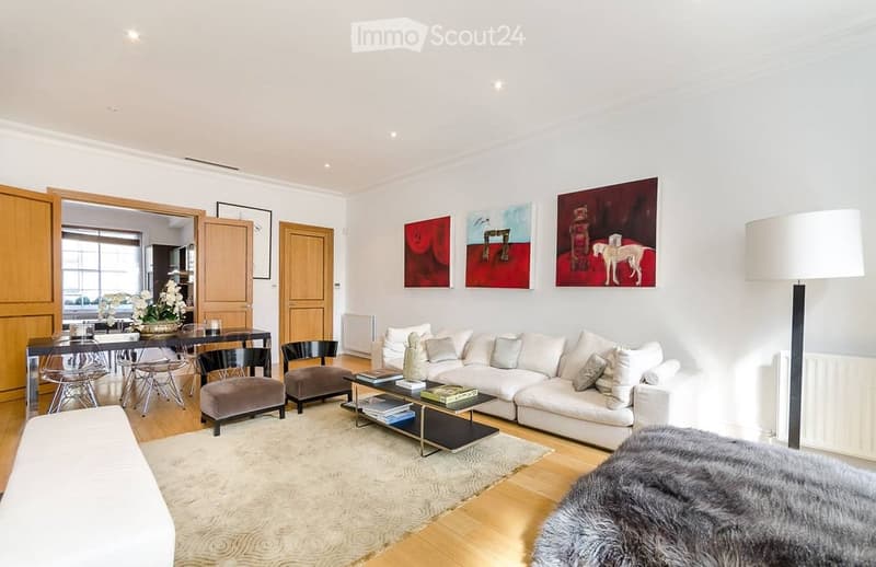 Modern living room with white walls, wooden flooring, a large white sectional sofa, a coffee table, two ottomans, a dining table with chairs, and artwork on the walls.