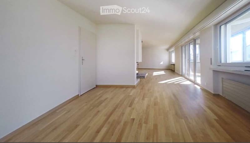 Empty room with wooden flooring, white walls, large windows, and a door leading to another area.