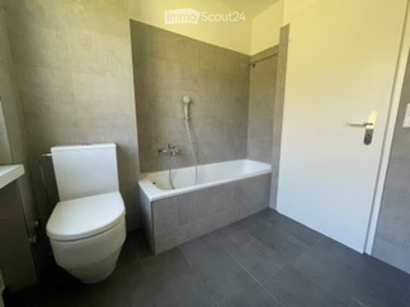 Modern bathroom with a white toilet, a rectangular bathtub, and a shower head attached to the wall. The floor is dark grey tiles, and the walls are a mix of grey and white tiles.