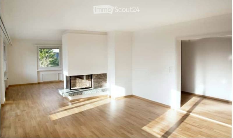 Empty room with light wooden flooring, white walls, and a central fireplace. Large window on one side allows natural light to enter.