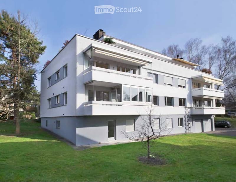Modern white multi-story building with multiple balconies and large windows, surrounded by a well-maintained lawn and trees.
