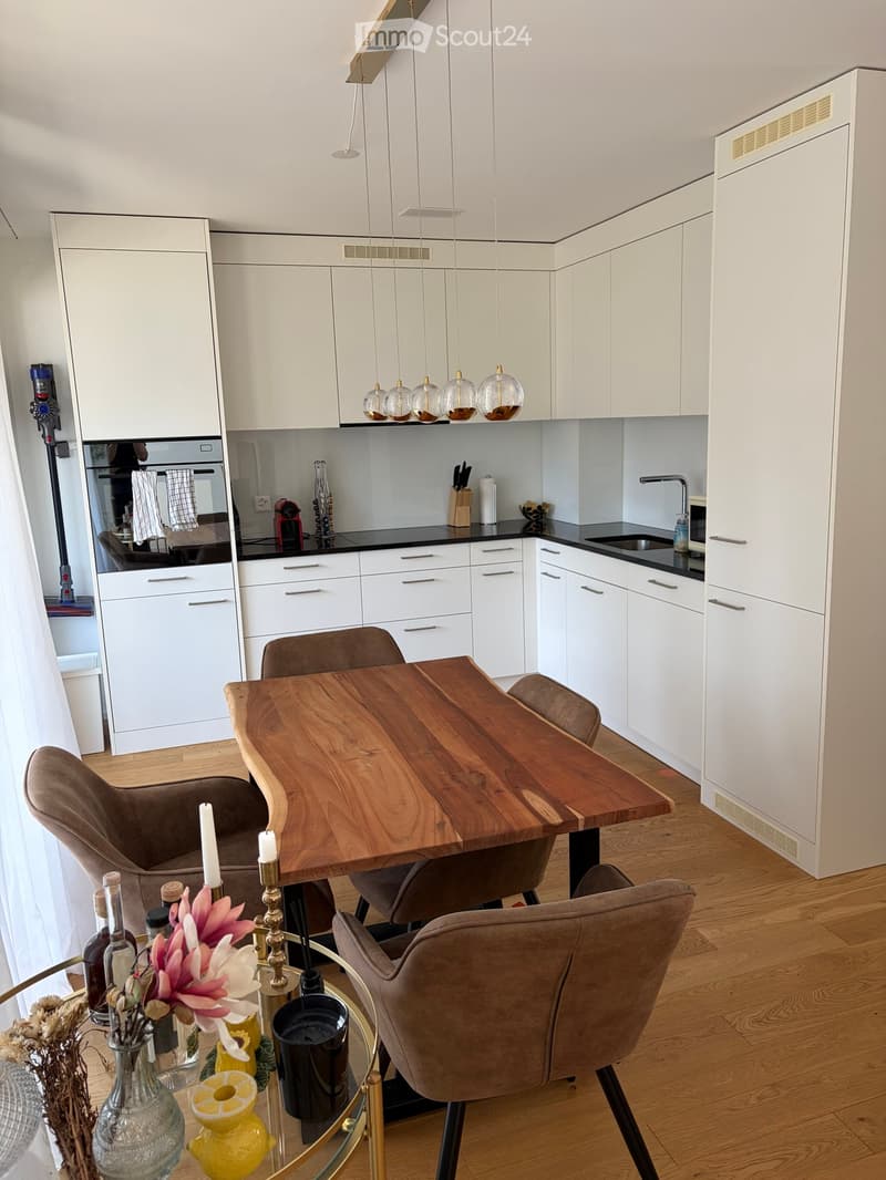 Modern kitchen with white cabinets, a wooden dining table, and pendant lights. Includes a sink, faucet, and various decorative items on the counter.