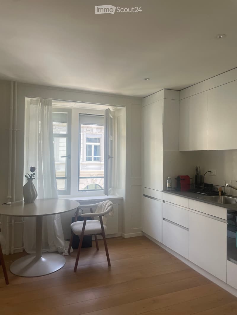 Bright, modern room with a round table, white chair, vase, and kitchen area with cabinets, sink, and oven.