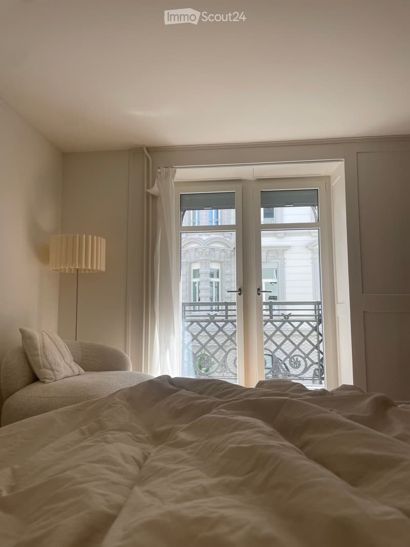 Cozy bedroom with a large bed, pillows, a sofa, and a floor lamp near a large window with a view of a balcony.