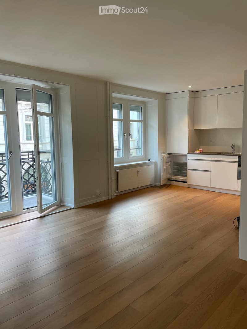 Empty room with light wooden flooring, white walls, large windows, and a small kitchen area with white cabinets and a sink.