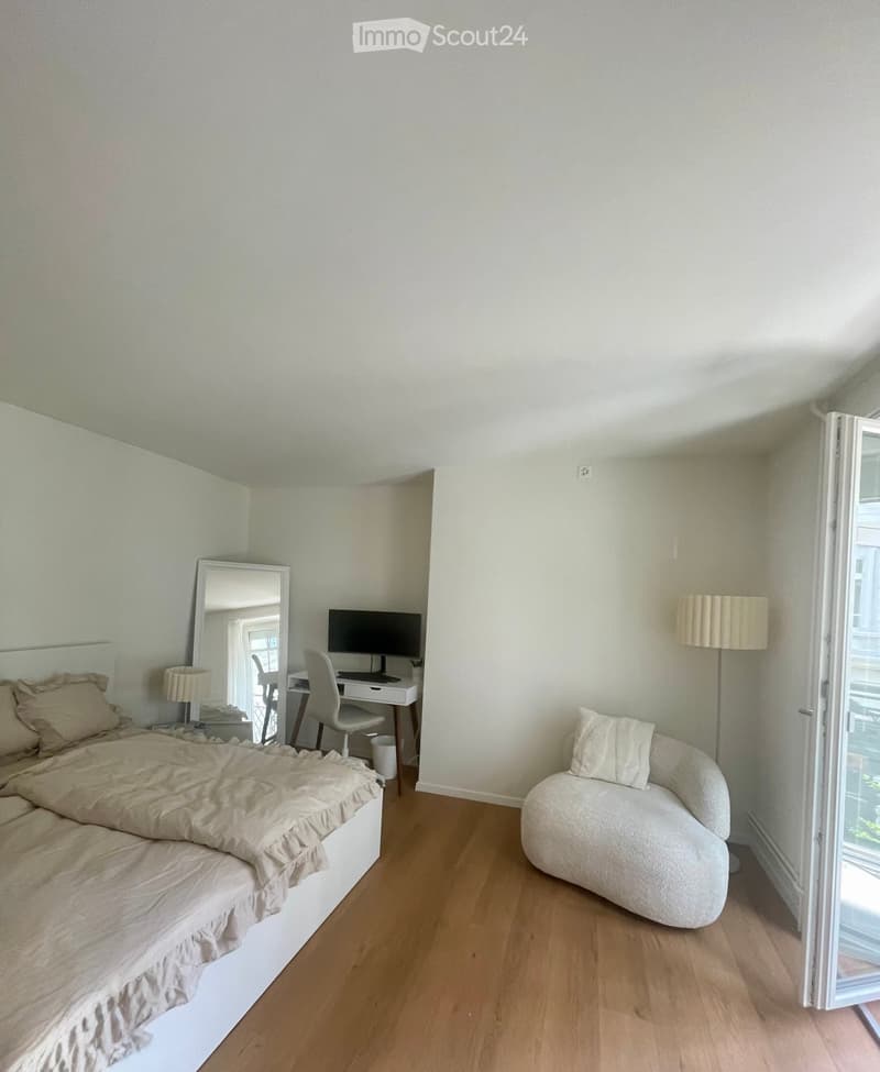 Cozy bedroom with a bed, sofa, desk, chair, computer, mirror, and lamps. The room features light-colored walls and wooden flooring.