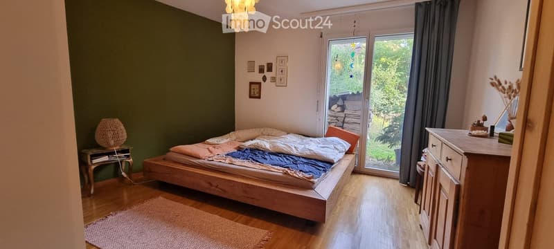 Cozy bedroom with wooden floor, large bed with pillows and blankets, nightstand with lamp, cabinet, and a view of the garden through large windows.