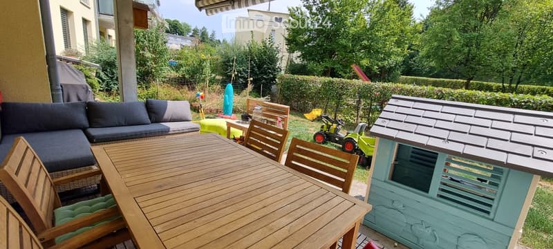 Outdoor patio area with a wooden dining table, chairs, a sofa, and a small playhouse. The space is surrounded by greenery and includes a covered seating area with a canopy.