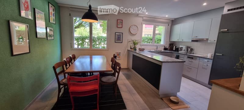 Modern kitchen with a central island, dining table with chairs, and various appliances. The space features light-colored cabinets, a granite countertop, and large windows with a view of greenery.