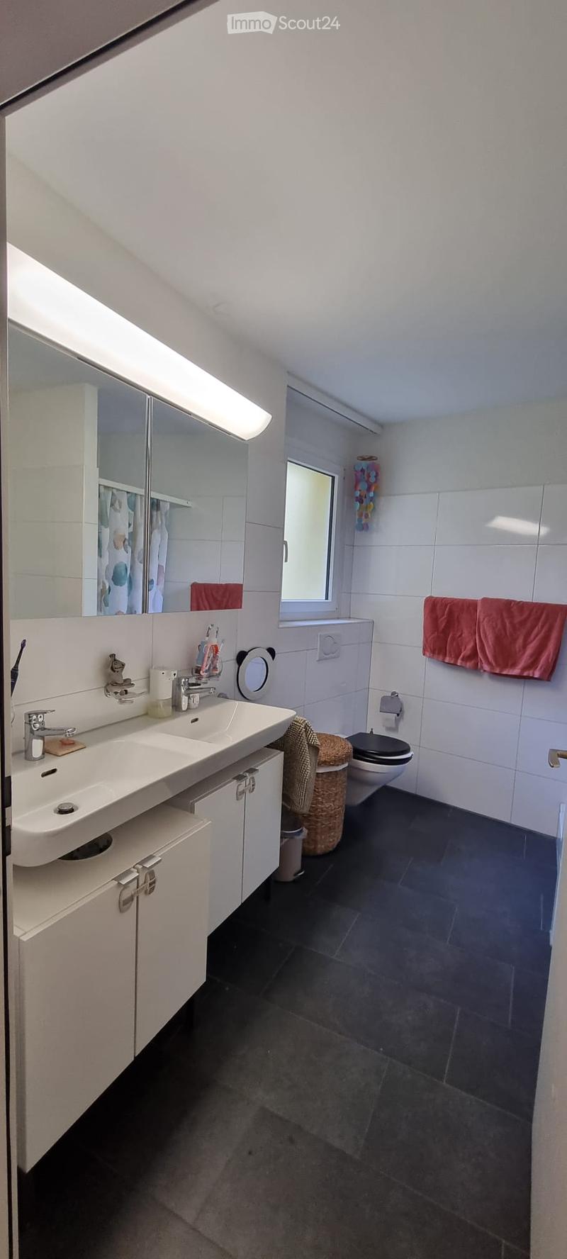 Modern bathroom with white tiles, dark floor, double sink, mirror, toilet, and red towels.