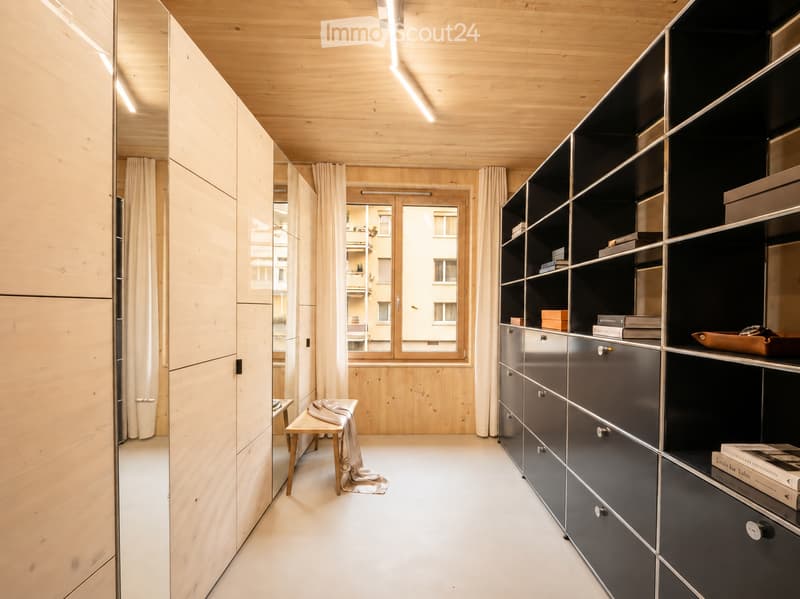 Modern hallway with light wooden walls, white floor, built-in dark grey cabinets, a small wooden bench with a cloth draped over it, and a large window with curtains leading to a balcony.